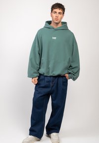 Ung man i en turkos oversized hoodie, baggy mörkblå jeans och vita sneakers, stående mot en enfärgad ljus bakgrund.