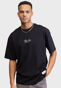 Ung mand iført en sort oversized T-shirt med teksten "Yee Haw", guld kædehalskæde og hånden i lommen foran en lys ensfarvet baggrund.