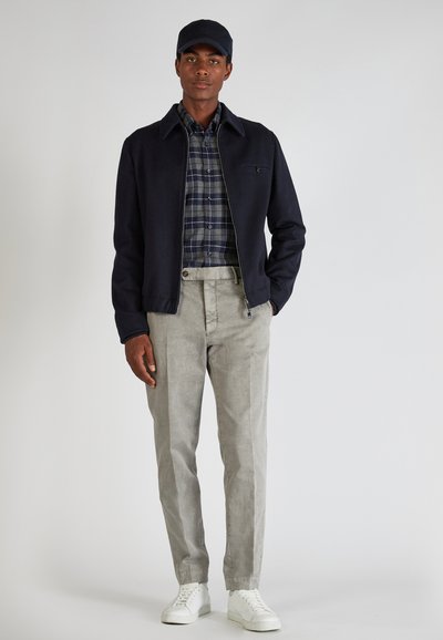 Chaqueta bomber navy sobre una camisa de cuadros, pantalones gris claro y zapatillas blancas, con una gorra oscura que complementa el conjunto.