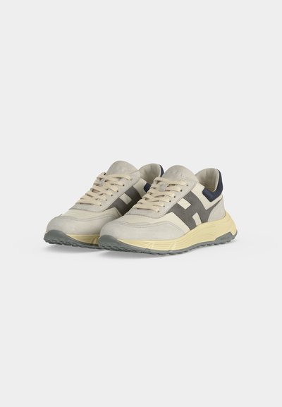 Paire de baskets beiges et grises à lacets avec des accents foncés et des semelles blanc cassé sur un fond clair uni.