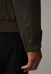 Brauner Jackenärmel mit geripptem Saum und strukturiertem Bündchen. Mit Druckknopfdetails und glattem Stoff. Sichtbare Nähte entlang der Naht.