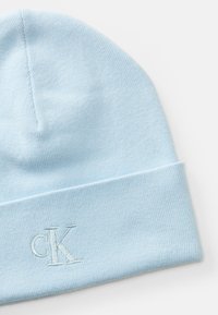 Lichtblauwe gebreide beanie met omgeslagen rand en geborduurd "cK" logo aan de voorkant, platgelegd op een witte achtergrond.