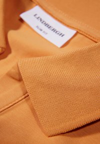 Tessuto arancione con un primo piano di un colletto e un'etichetta che recita "LINDENBURGH Slim Fit." Il materiale è testurizzato con una finitura liscia.