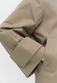 Beige takki, jonka hihansuu on taitettu, suunniteltu terävällä siluetilla. Ominaisuuksiin kuuluu sileä, teksturoidusta kankaasta valmistettu materiaali ja sivutasku.