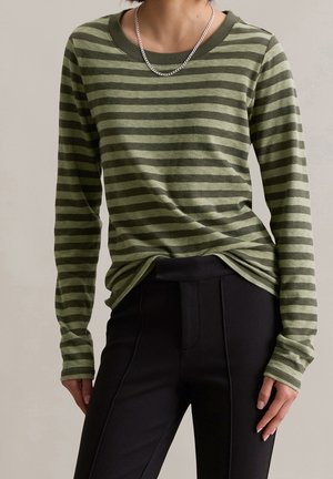 Person trägt ein grünes und dunkelgrün gestreiftes Langarmshirt mit einer silbernen Kettenhalskette und schwarze, eng anliegende Hosen vor einem einfarbigen Hintergrund.