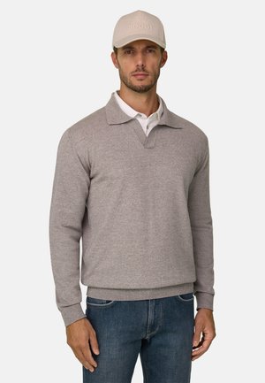 Homme portant une casquette beige, un pull gris clair à manches longues par-dessus une chemise blanche à col, et un jean bleu, debout devant un fond uni.
