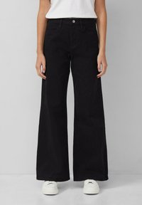 Schwarze weit geschnittene Jeans aus Baumwolle, mit hoher Taille, Fronttaschen und einem einzelnen Knopfverschluss. Kombiniert mit weißen Sneakern.