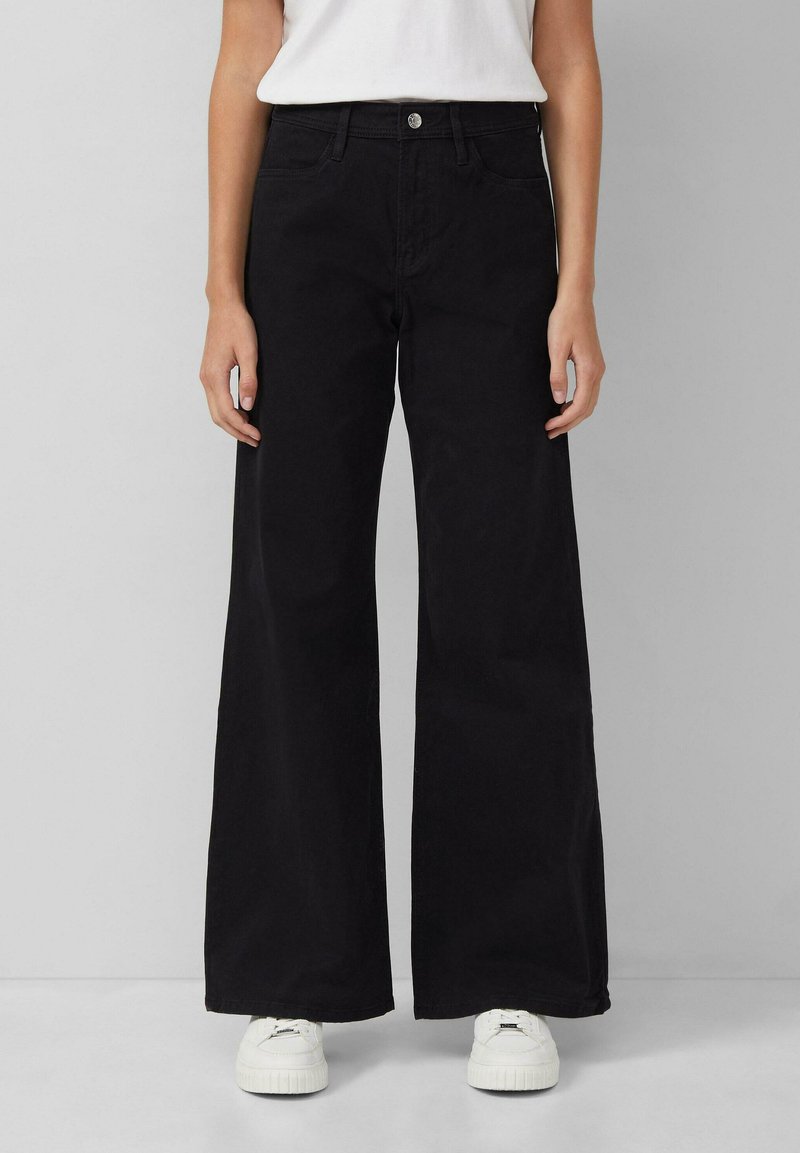 Schwarze weit geschnittene Jeans aus Baumwolle, mit hoher Taille, Fronttaschen und einem einzelnen Knopfverschluss. Kombiniert mit weißen Sneakern.