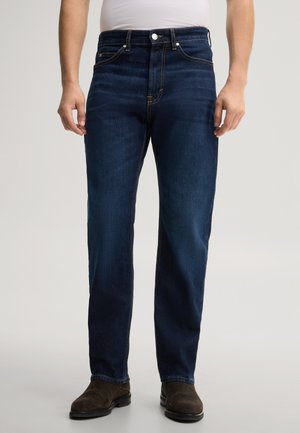 Mann, der dunkelblaue Jeans mit geradem Bein und braune Schuhe trägt, steht mit entspannten Armen an den Seiten vor schlichtem Hintergrund.