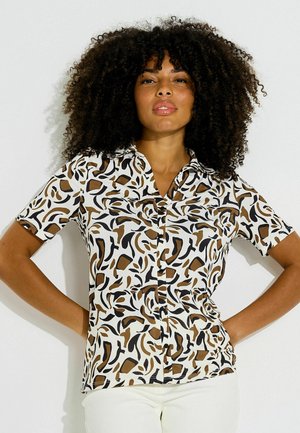 Femme aux cheveux bouclés portant une chemise à manches courtes boutonnée avec des motifs abstraits noirs et marron et un pantalon blanc, debout les mains sur les hanches.