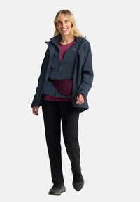 Giacca impermeabile blu navale con cappuccio, indossata sopra una camicia bordeaux e pantaloni scuri. Tessuto texture, cerniera frontale e tasche laterali visibili.