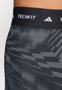 Sort elastisk taljebånd med hvid "TECHFIT" tekst. Mørkegrå tætsiddende stof med et geometrisk mønster og glat tekstur.