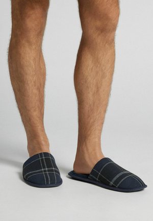 Marine ternede hjemmesko med en blød stofoverdel og polstret sål. Har et slip-on design og et gråt ternet mønster for ekstra detaljer.