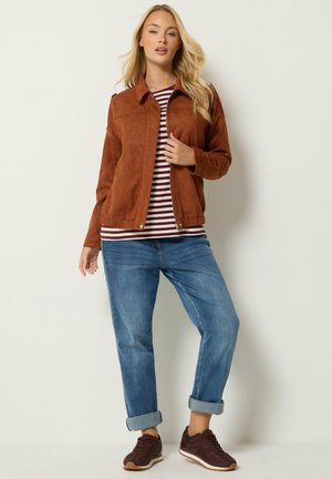 Donna che indossa una giacca in camoscio marrone sopra una camicia a righe, jeans blu risvoltati e sneakers marroni, in piedi davanti a un muro chiaro e semplice.