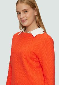 Pull orange à texture avec un motif matelassé et une chemise à col blanc en dessous, mettant en valeur un décolleté en rond et des manches longues.