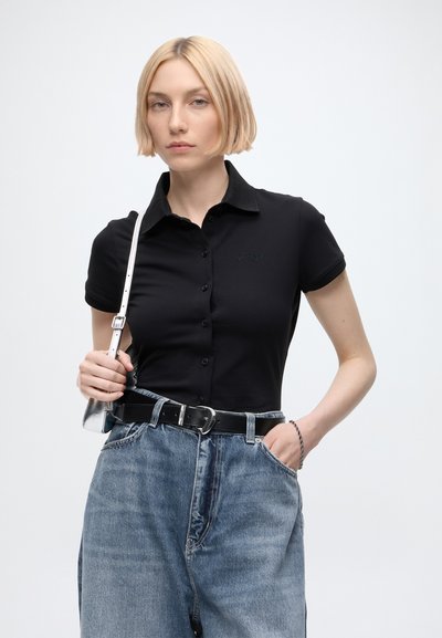 Jeune femme aux cheveux blonds courts portant un polo noir boutonné, un jean bleu avec une ceinture noire, et tenant une lanière de sac à main argentée.