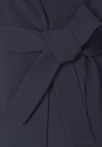 Dettaglio di un vestito avvolgente blu navy con una fascia in tessuto annodata a fiocco, caratterizzato da una superficie liscia e un leggero lucido. Include tasche laterali.