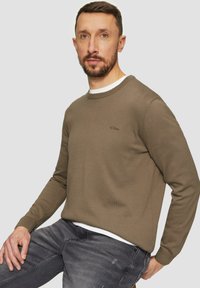 Man met kort bruin haar en een baard, gekleed in een olijfgroene trui over een wit shirt en donkergrijze jeans, zittend en vooruitkijkend.