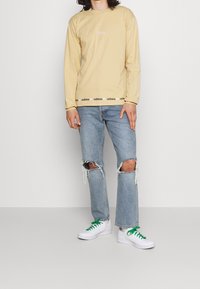 Långärmad beige t-shirt med liten logotyp, tillsammans med ljusblå slitna jeans. Vita sneakers med gröna snören.