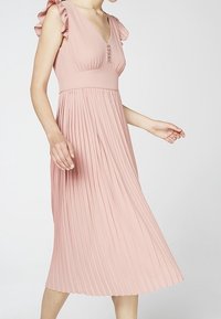 Eine Frau trägt ein ärmelloses, hellrosa gefältetes Midikleid mit Rüschen an den Schultern und kleinen Knöpfen am V-Ausschnitt.