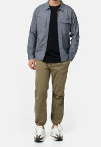 Homme portant une veste grise zippée, un t-shirt noir, un pantalon cargo beige avec des poignets élastiques et des baskets blanches, debout devant un fond uni.