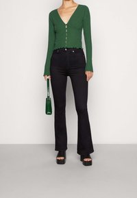 Grünes, langärmliges, V-ausschnitt-rippstrick Crop Top mit Knopfverschluss. Hoch taillierte schwarze Schlagjeans, schwarze Plateauschuhe und eine kleine grüne Handtasche.