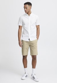 Chemise blanche à manches courtes avec boutons, shorts kaki, baskets blanches avec des rayures noires et chaussettes blanches. Tenue d'été simple et décontractée.