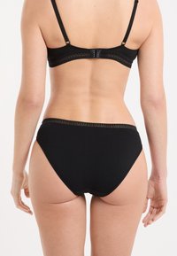 Donna vista di spalle che indossa un reggiseno nero e slip abbinati con finiture in pizzo su sfondo bianco.