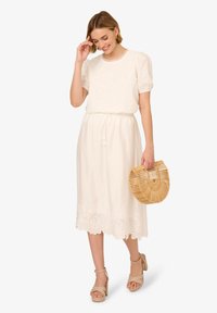 Abito bianco con ricami floreali all'orlo, maniche a sbuffo corte, vita aderente. La modella tiene in mano una borsa rotonda in vimini, indossando tacchi a piattaforma beige.