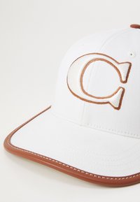 White cap featuring a prominent stylized letter "C" in brown embroidery. Brown trim along the brim, textured fabric.