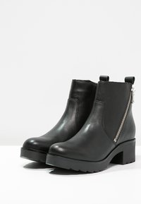 Botas de cuero negro hasta el tobillo con un tacón grueso, que cuentan con una cremallera lateral y una textura suave, diseñadas para facilitar el uso y el estilo.