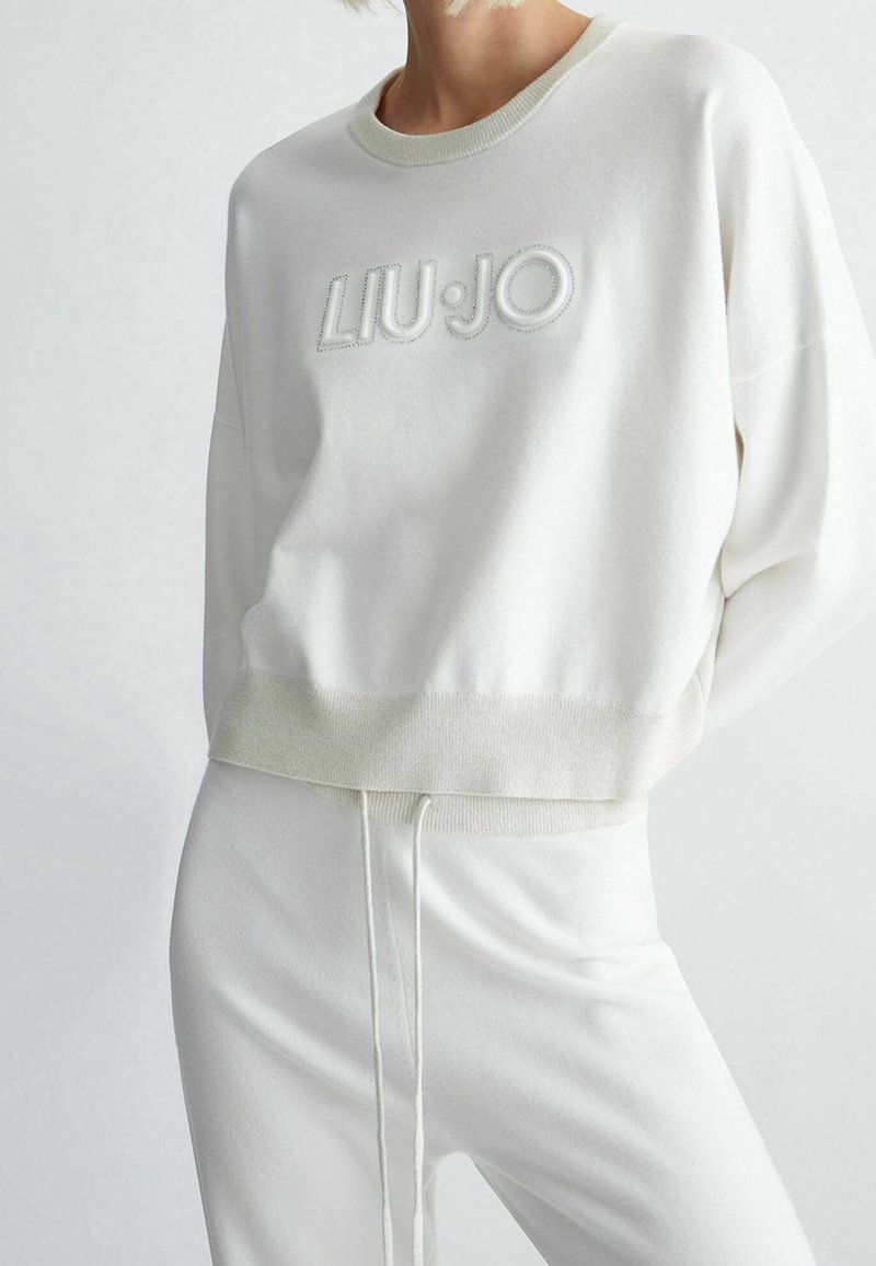 Femme portant un sweat-shirt court blanc avec le logo "LIU·JO" et un pantalon blanc taille haute assorti avec une taille à cordon.