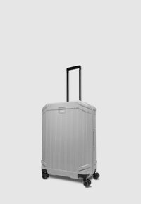 Maleta rígida gris con estrías verticales, asa negra retráctil y cuatro ruedas de giro suave. Incluye bolsillo lateral y etiqueta con el logo.