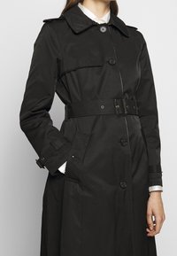 Trench-coat noir en tissu résistant à l'eau. Doté d'une taille ceinturée, de grands boutons à l'avant et d'un col plié. Design minimaliste.