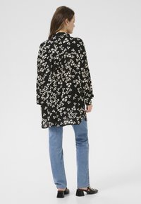 Langärmliges schwarzes Hemd mit floralem Muster, leichtes Material, lockere Passform und gefaltetem Rückenteil. Kombiniert mit blauen Jeans und schwarzen Schuhen.