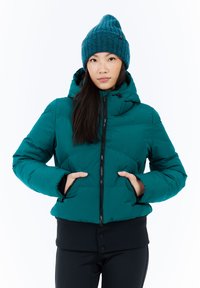 Moteris, vilkinti teal spalvos pūkinę striukę su kapišonu ir suderinta megzta kepure, rankomis kišenėse, stovinti prieš baltą fono.
