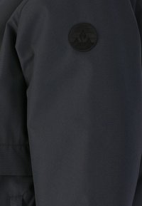 Giacca nera, resistente all'acqua, con una texture di tessuto liscia, polsini elastici e un logo a toppa rotondo con grafica di montagne sulla spalla.