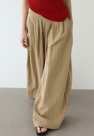 Donna con pantaloni beige a gamba larga, top rosso senza maniche e sandali neri, in piedi su pavimento grigio davanti a una parete chiara.