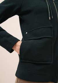 Tmavě zelená zipová bunda s vroubkovanou texturou, obsahující velkou přední kapsu se zapínáním na patentku. Jsou zde také nenápadné stahovací šňůrky.