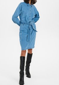 Denimskjortklänning i ljusblått med bälte i midjan, långärmade, knäppning med knappar, och knälång kjol. Matchad med knähöga svarta stövlar.