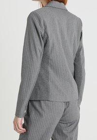 Donna che indossa un blazer grigio a righe sottili aderente e pantaloni coordinati, mostrata di spalle su uno sfondo semplice.