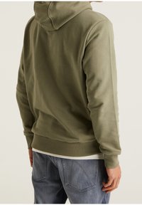 Felpa verde oliva con una finitura testurizzata, dotata di cappuccio con coulisse e polsini a coste. Abbinata a jeans in denim azzurro chiaro.