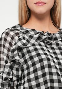 Blouse à carreaux noir et blanc avec un col volanté, en tissu léger, présentant des carrés de tailles variées et une surface texturée.