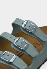 Τρεις λουρίδες από δέρμα σε ανοιχτό γκρι χρώμα με ρυθμιζόμενες ασημένιες μεταλλικές αγκράφες πάνω σε καφέ πέλμα.