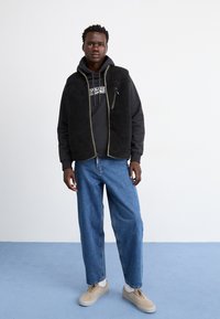 Gilet en polaire noir avec des bords verts porté sur un pull à capuche noir et un jean bleu ample. Le mannequin portant des baskets beiges se tient sur un fond bleu clair.