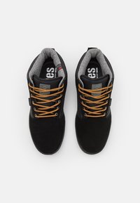 Sapatos de camurça pretos com forro de lã cinza, atacadores laranja e colarinho acolchoado; possuem uma sola de borracha preta e logotipo na língua.