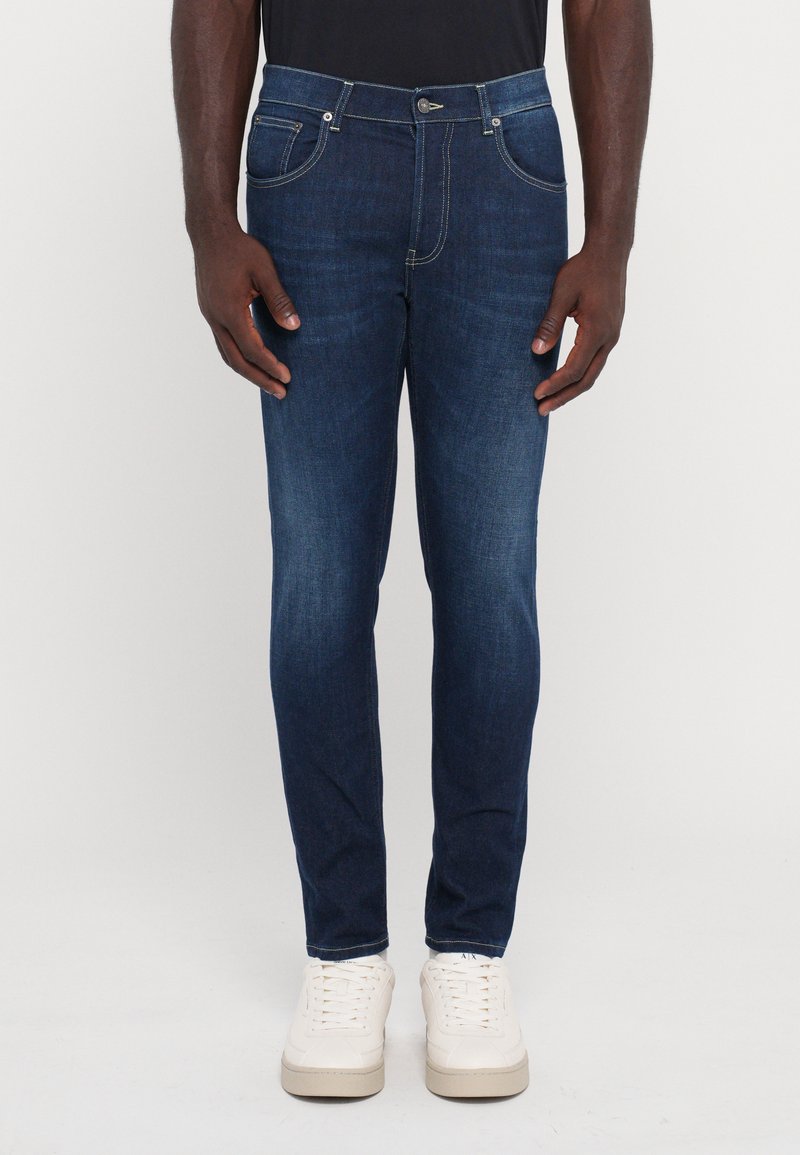 Jeans din denim de un albastru închis, cu croi slim, având cinci buzunare, cusături galbene și un nasture cu prindere metalică.