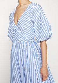 Femme portant une robe portefeuille rayée verticale bleu et blanc avec des manches ballon aux coudes et un profond décolleté en V.