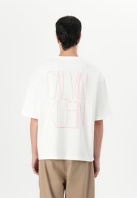 Bílý bavlněný tričko s velkým růžov�ým logem "CALVIN KLEIN" na zadní straně. Volný střih s krátkými rukávy a kulatým výstřihem.