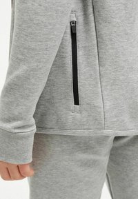 Grauer Sweatshirt mit glatter Textur, ausgestattet mit einer schwarzen Reißverschlusstasche und einem Bündchen am Handgelenk, kombiniert mit passenden grauen Hosen.